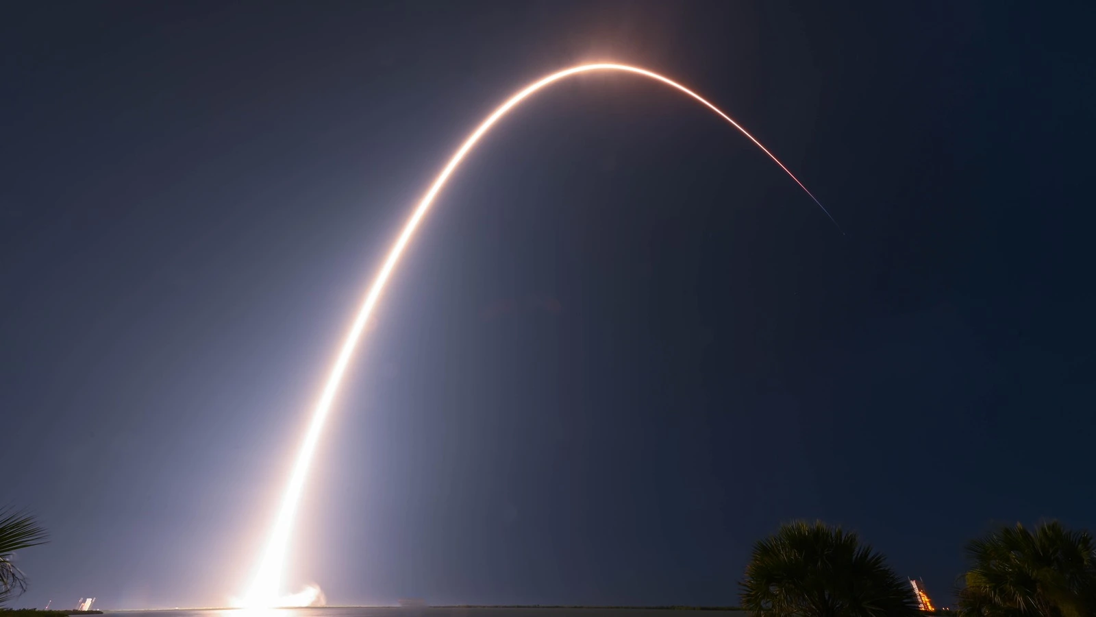 Long-exposure photo of a rocket launch arcing over water and palm trees at night, Cape Canaveral, Florida.